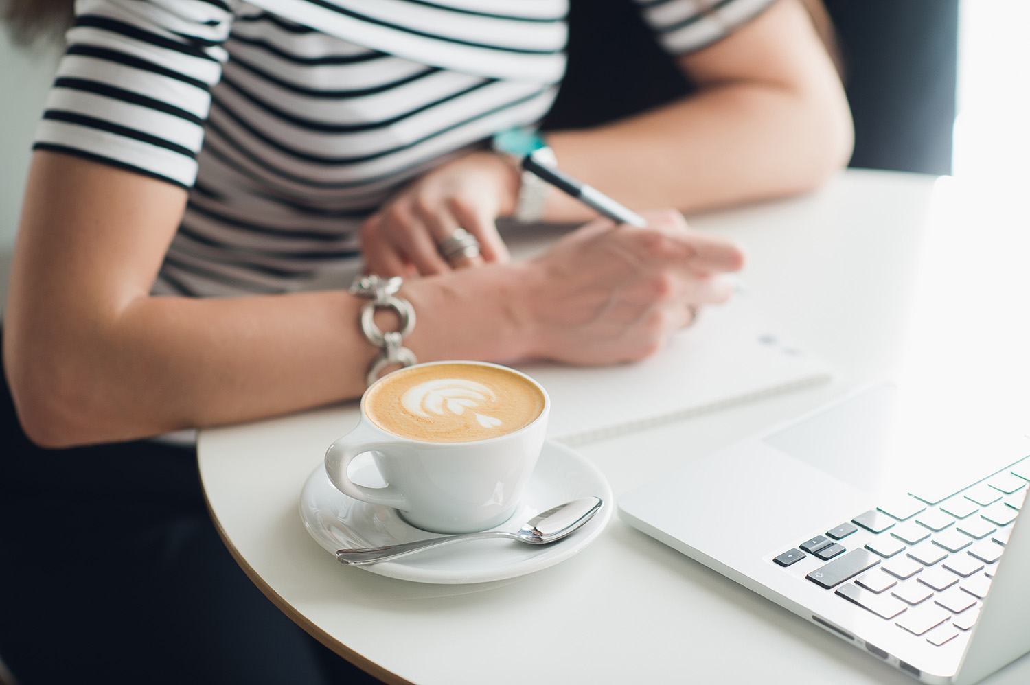zdjęcie osoby piszącej w notatniku przed otwartym laptopem, z boku filiżanka kawy
